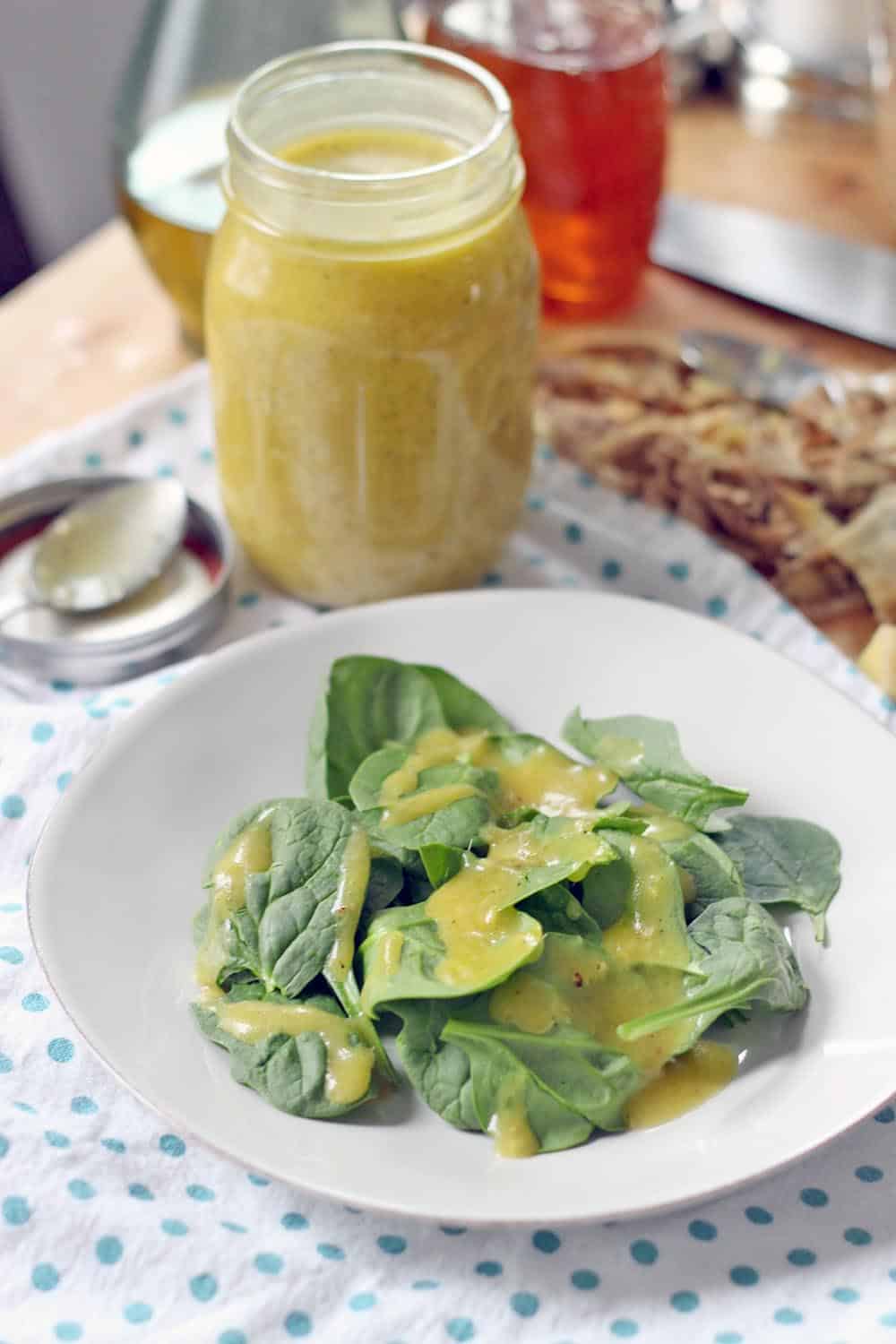 White plate holding greens drizzled with Fresh Ginger and Honey Mustard Dressing Marinade. A glass jar filled with the dressing/marinade, as well as ingredients, are in the background.