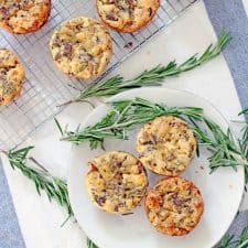 Bird's eye view of a white plate holding three mini quiches, next to a wire cooling rack holding several more mini quickes. Sprigs of rosemary are scattered around.