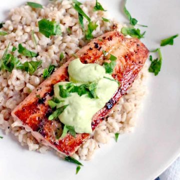 Bird's eye view of a white plate holding a salmon filet on top of a bed of brown rice, with a dollop of Avocado Lemon and Garlic Aioli on the sauce. The meal is sprinkled with chopped herbs.