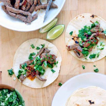 Bird's eye view of a wooden cutting board holding two open tacos, a plate of tortillas, a plate of steak, lime wedges, and chopped greens.