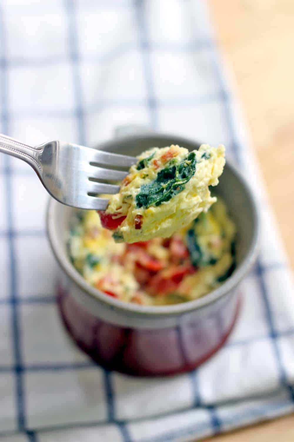 A fork lifts a bite of Five Minute Spinach and Cheddar Microwave Quiche in a Mug up from a blurred-out mug.