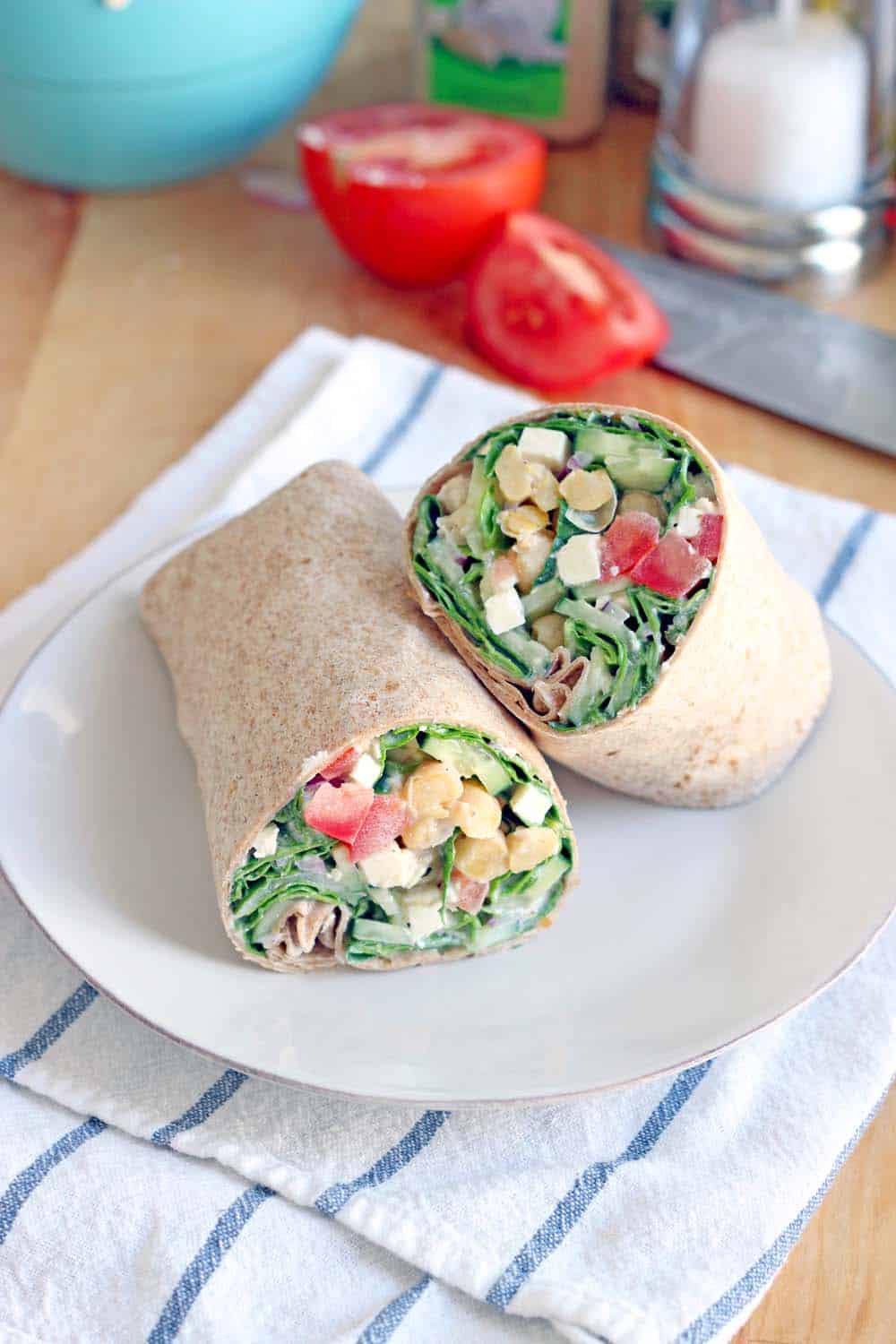 Two halves of a wrap filled with greens, chickpeas, tomatoes, feta, and other fillings, displayed on a white plate, on a blue and white striped cloth. Ingredients are in the background.