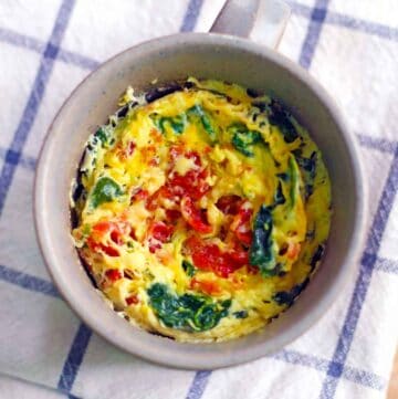 Square overhead photo of a microwave quiche in a mug with eggs, spinach, and tomato, on a blue checkered towel.