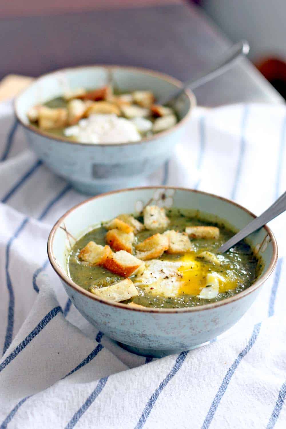 Two bowls of green soup topped with croutons and an egg; one in the foreground and one slightly blurred in the background.