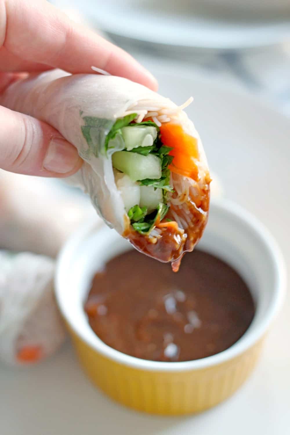 A light skinned hand holds a spring roll that has been cut in half up to the camera, so inside fillings are visible. The roll is partially dipped in peanut sauce and a ramekin of peanut sauce is blurred out in the background.