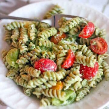 Square photo of a pesto pasta salad on a white plate with cherry tomatoes and a fork alongside.