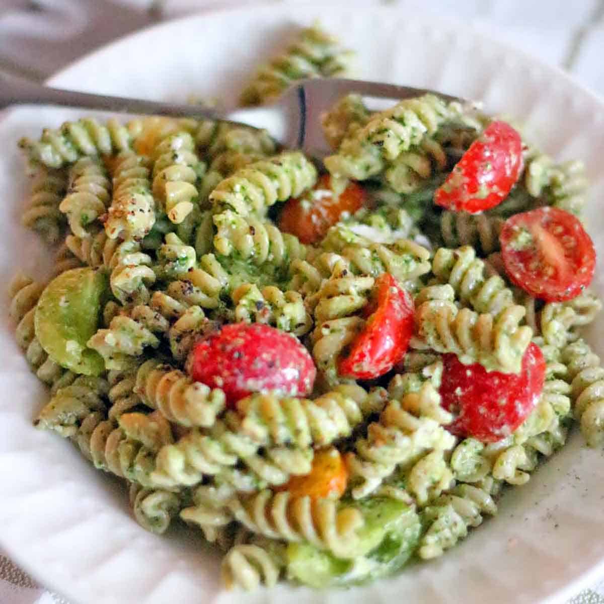 Square photo of a pesto pasta salad on a white plate with cherry tomatoes and a fork alongside.