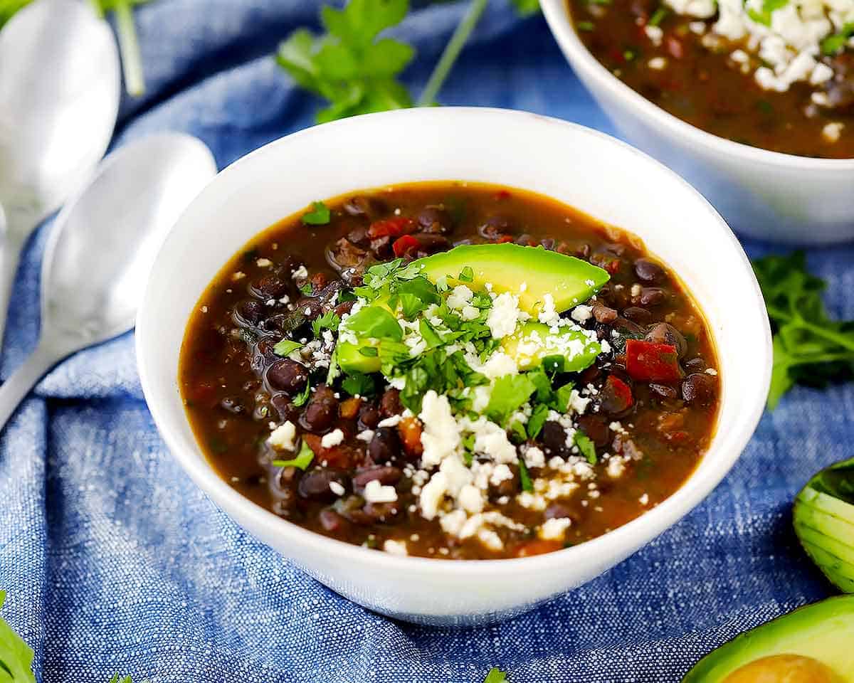 A bowl of black bean soup with avocado, cilantro, and cotija cheese on top on a blue towel.