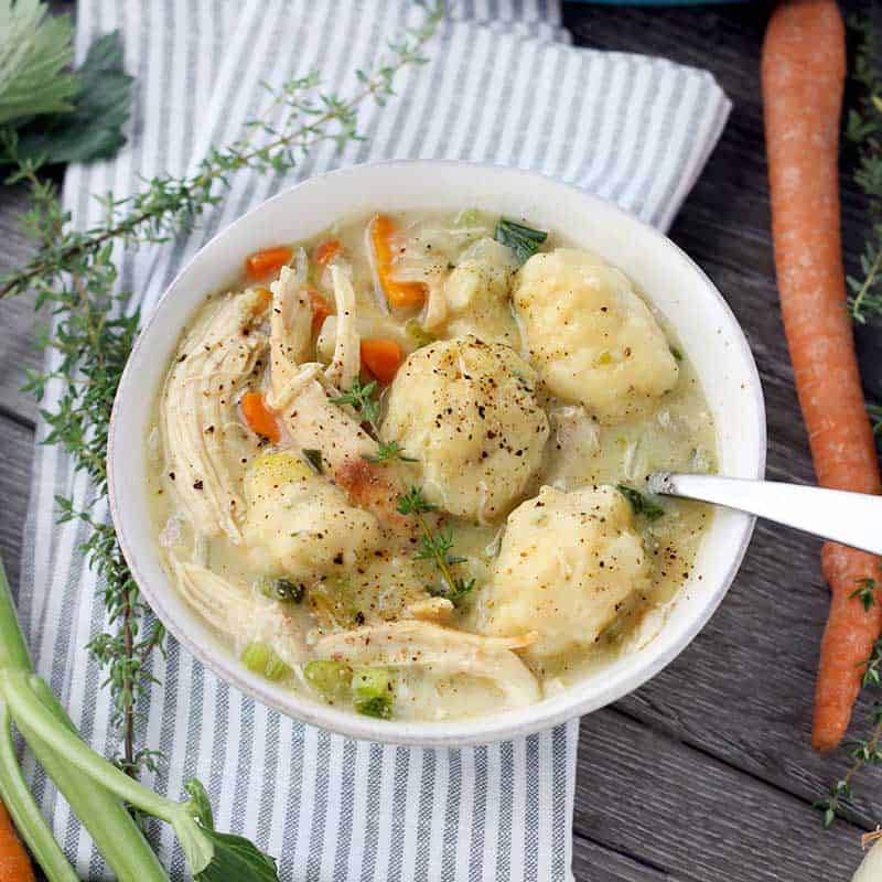 A bowl full of chicken and dumplings with a spoon and vegetables in the background