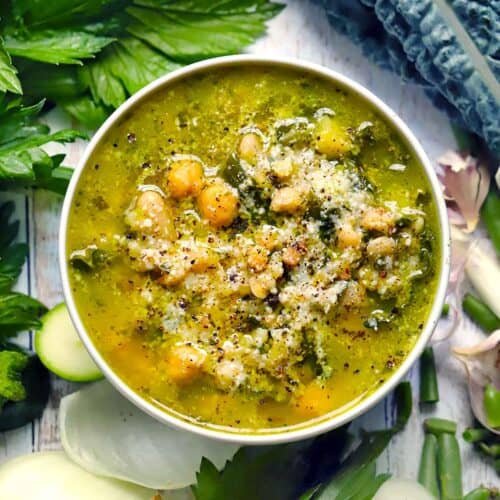 Square overhead photo of green pesto minestrone soup with chickpeas, white beans, and Parmesan in a white bowl, surrounded by fresh vegetables.
