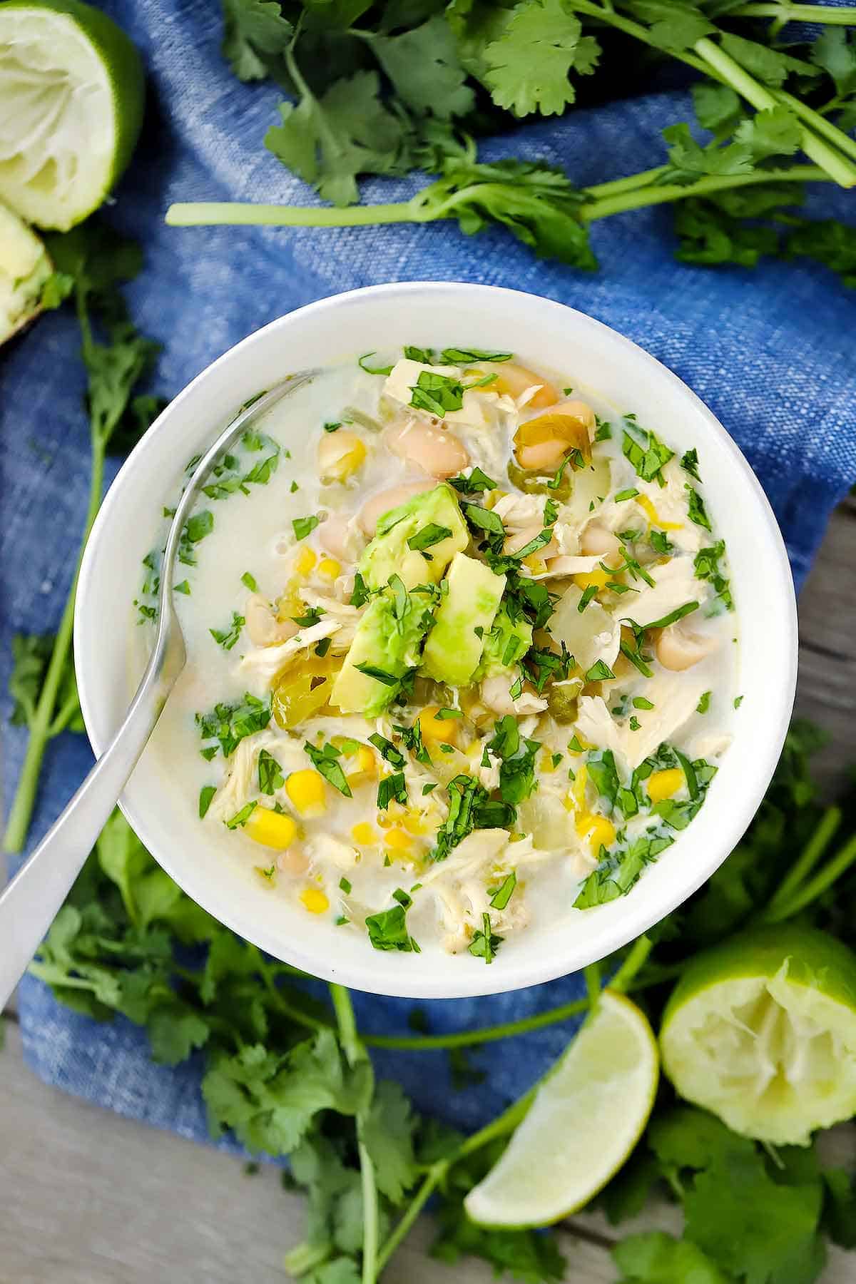 An overhead photo of a bowl of white chicken chili with cilantro and limes in the background on a blue towel.