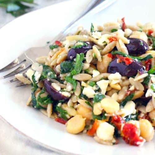 Square photo of a mediterranean chickpea orzo salad on a white plate with a fork alongside, with olives, vegetables, and herbs in the salad.