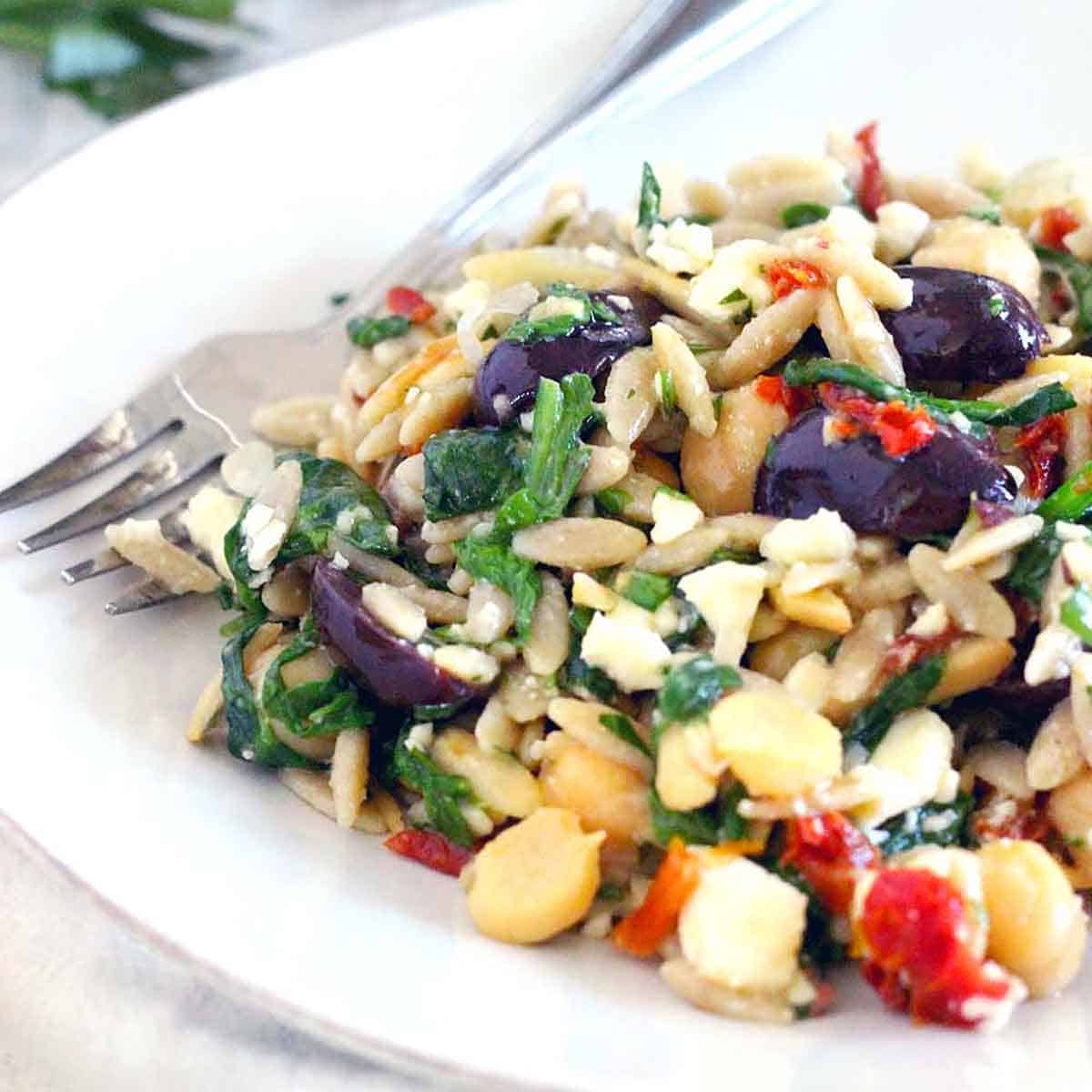 Square photo of a mediterranean chickpea orzo salad on a white plate with a fork alongside, with olives, vegetables, and herbs in the salad.
