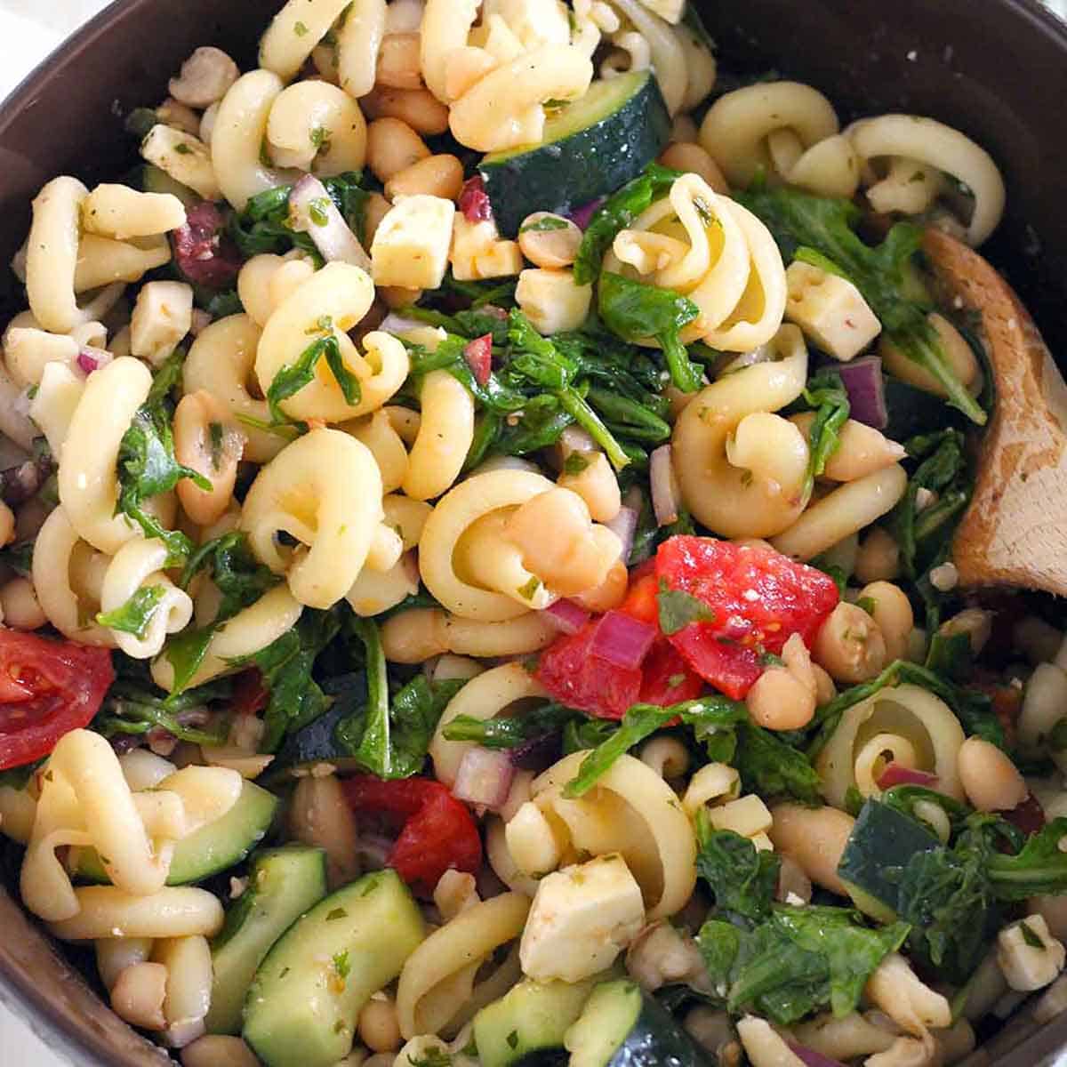 Square photo of close up overhead view of a Greek pasta salad with arugula, white beans, tomatoes, and herb vinaigrette.