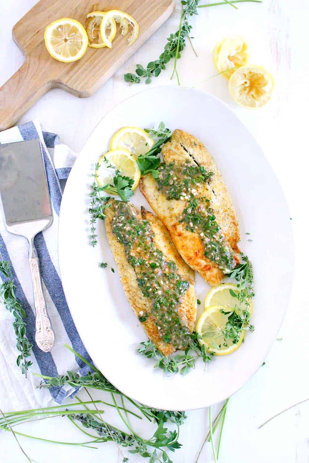 An overhead photograph of a platter of sea bass with lemon slices and a garlic herb sauce with a serving spatula.