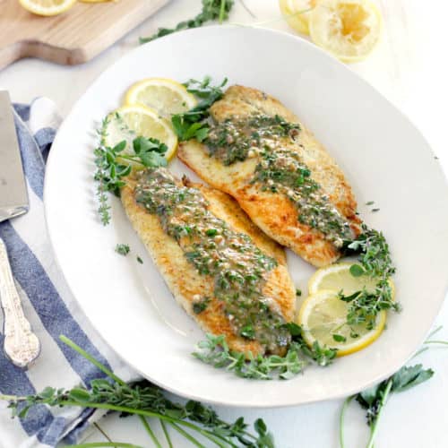 A platter of pan fried fish with herb sauce and lemon slices.