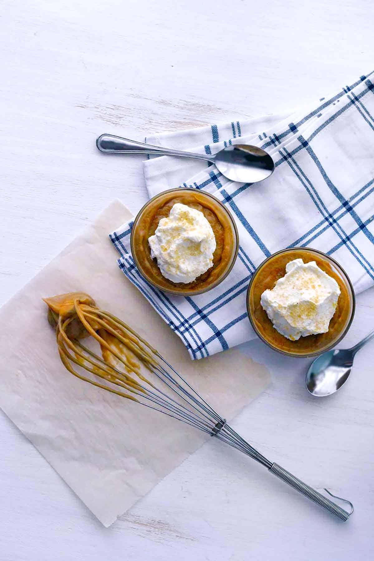 Two bowls of butterscotch pudding with a whisk covered in it on the side.