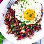 Square photo of corned beef hash on a plate with a fork and a fried egg on top.