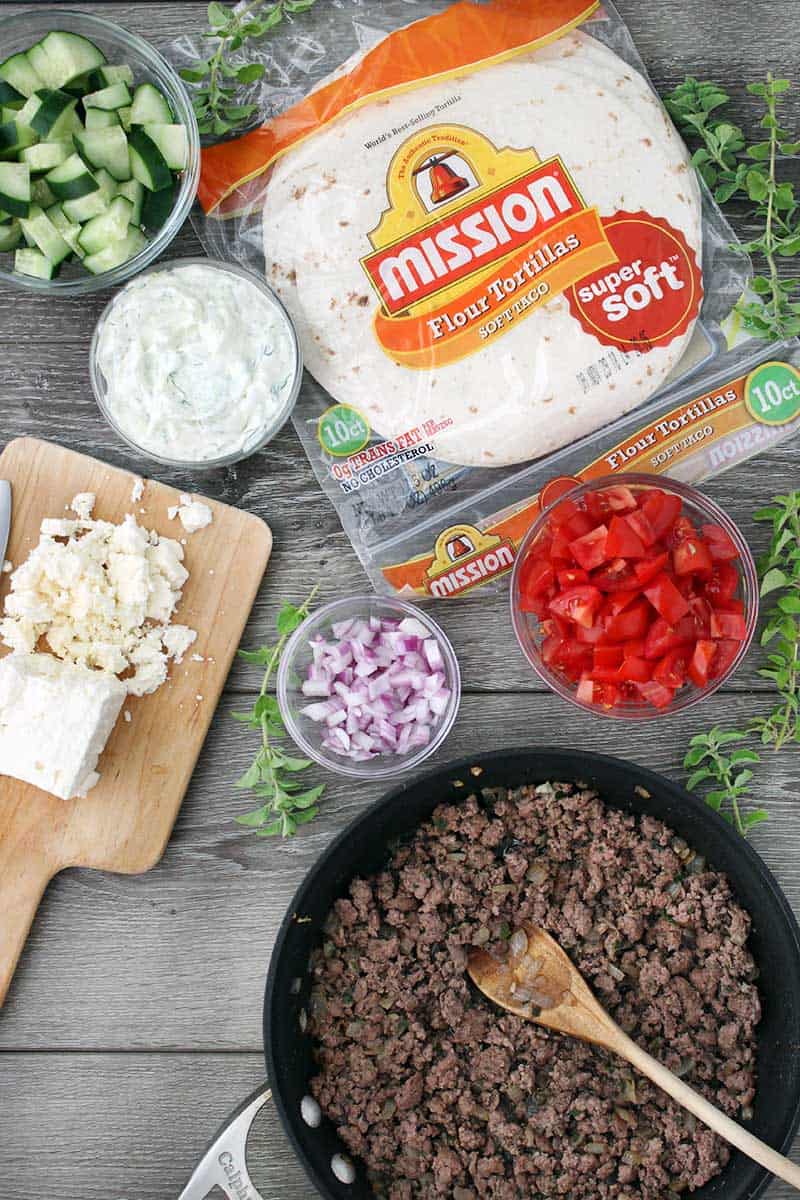 Mission flour tortillas, a skillet of ground lamb, red onions, cucumbers, feta cheese, and tzatziki sauce flatlay photo.