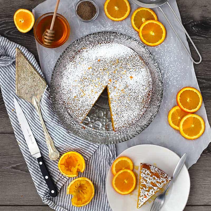 Square photo of an orange olive oil cake sprinkled with powdered sugar and orange slices around it.
