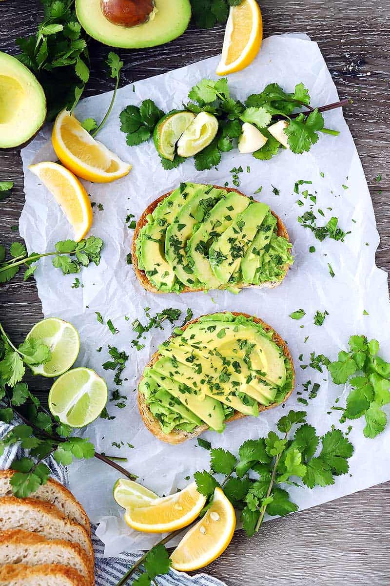 Overhead photo of avocado toast, cilantro, lemon and lime wedges, and bread.