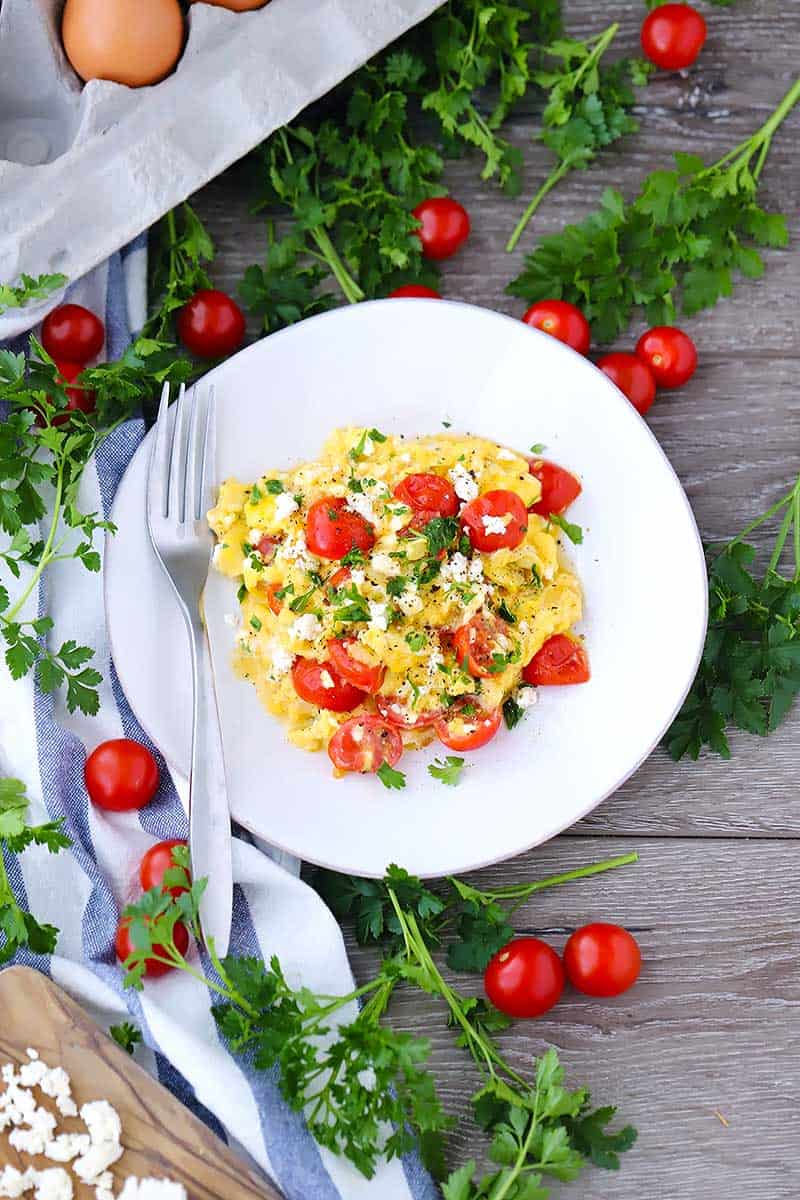 Overhead photo of scrambled eggs with feta and cherry tomatoes and parsley scattered around.