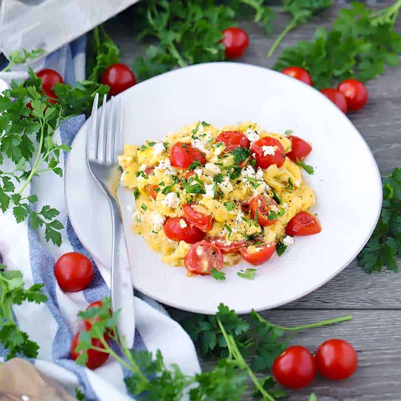 Square photo of a plate of scrambled eggs with a fork and tomatoes.