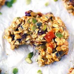 Square photo of a close up breakfast cookie made with oats, nuts, seeds, and dried fruit, with a bite taken out and pumpkin seeds scattered around.