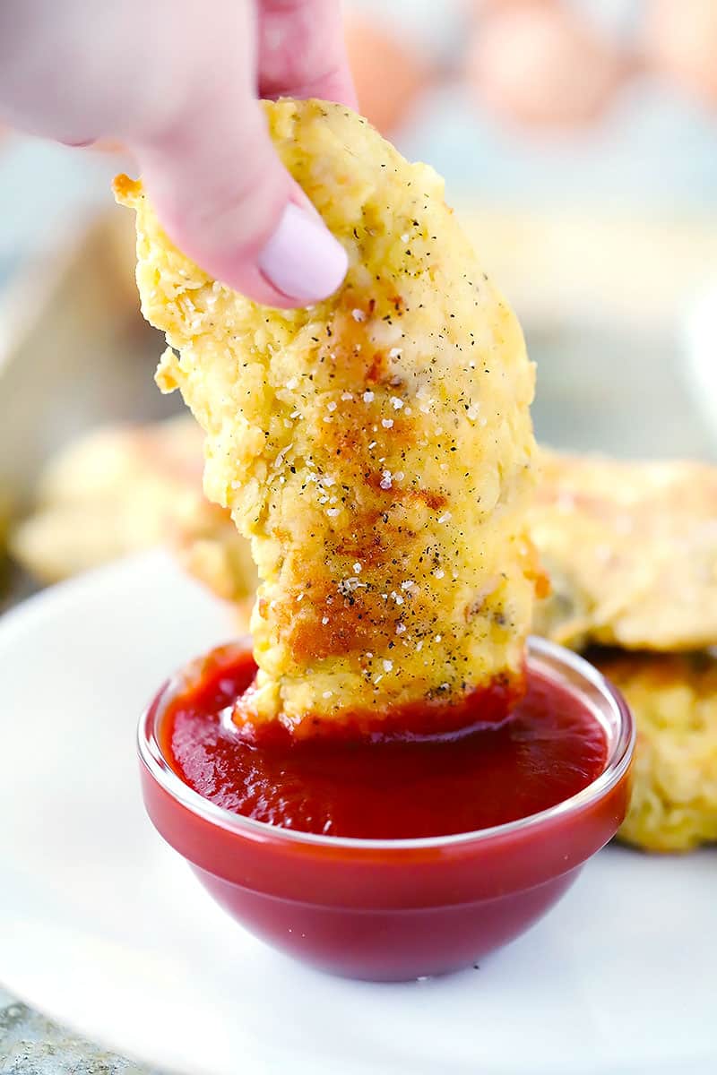 A hand holding a chicken tender and dipping it in ketchup.