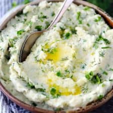 A bowl of mashed cauliflower with melted butter and fresh herbs and a spoon.