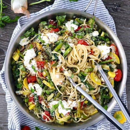 A skillet with spaghetti, vegetables, and burrata with tongs
