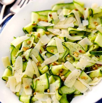 A close up photo of a zucchini ribbon salad with toasted pine nuts and shaved parmesan cheese on a white platter.