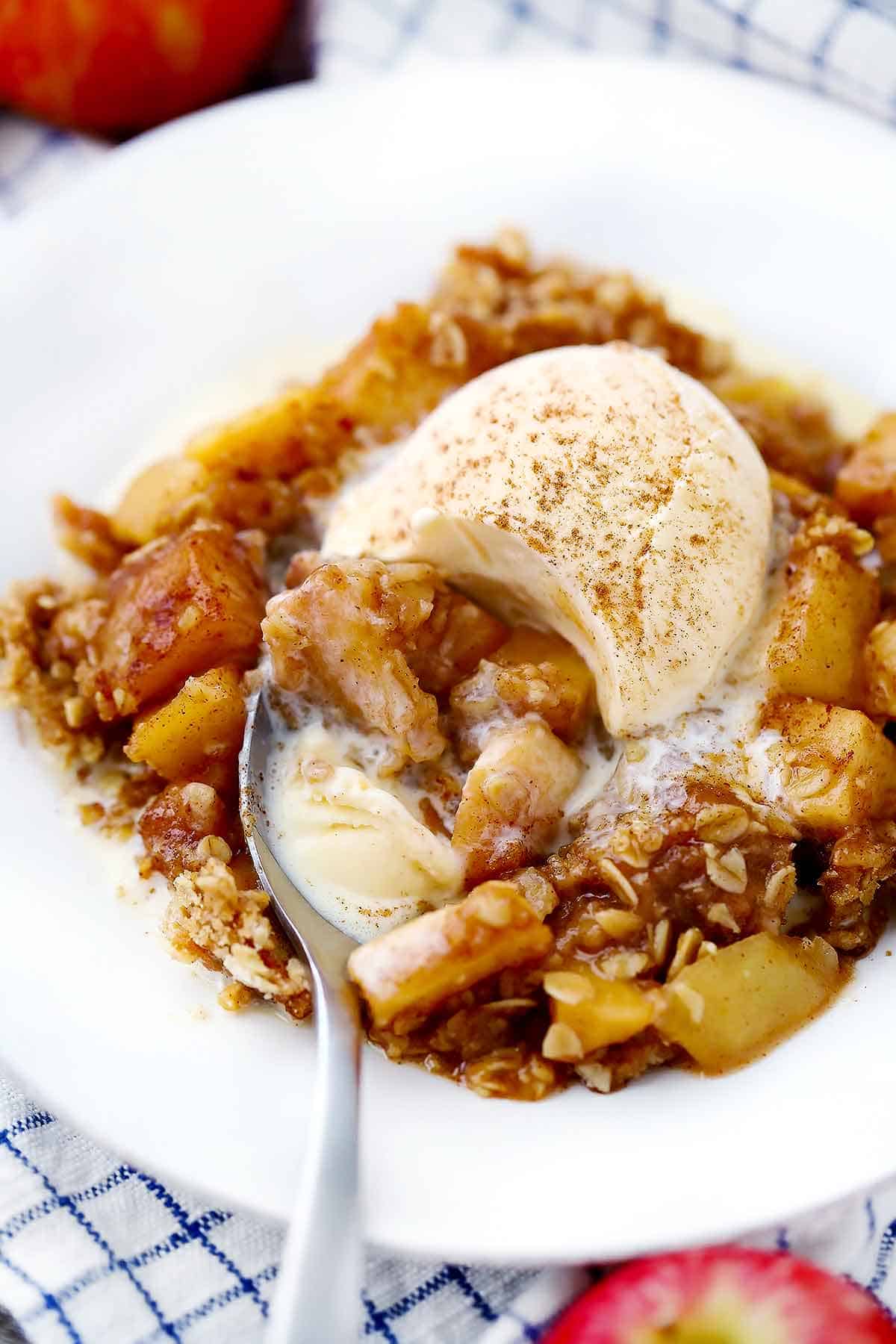 Apple crisp with vanilla ice cream melting on top and a spoon taking a bite out of it.