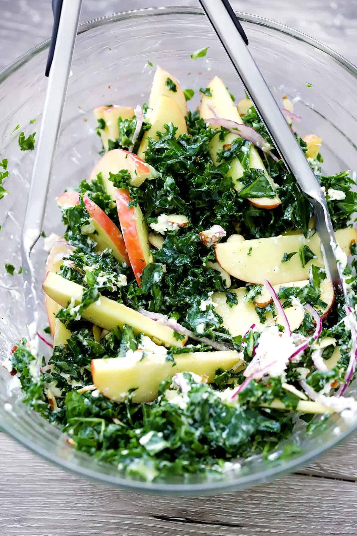 Mixing a kale salad together in a glass bowl with tongs.