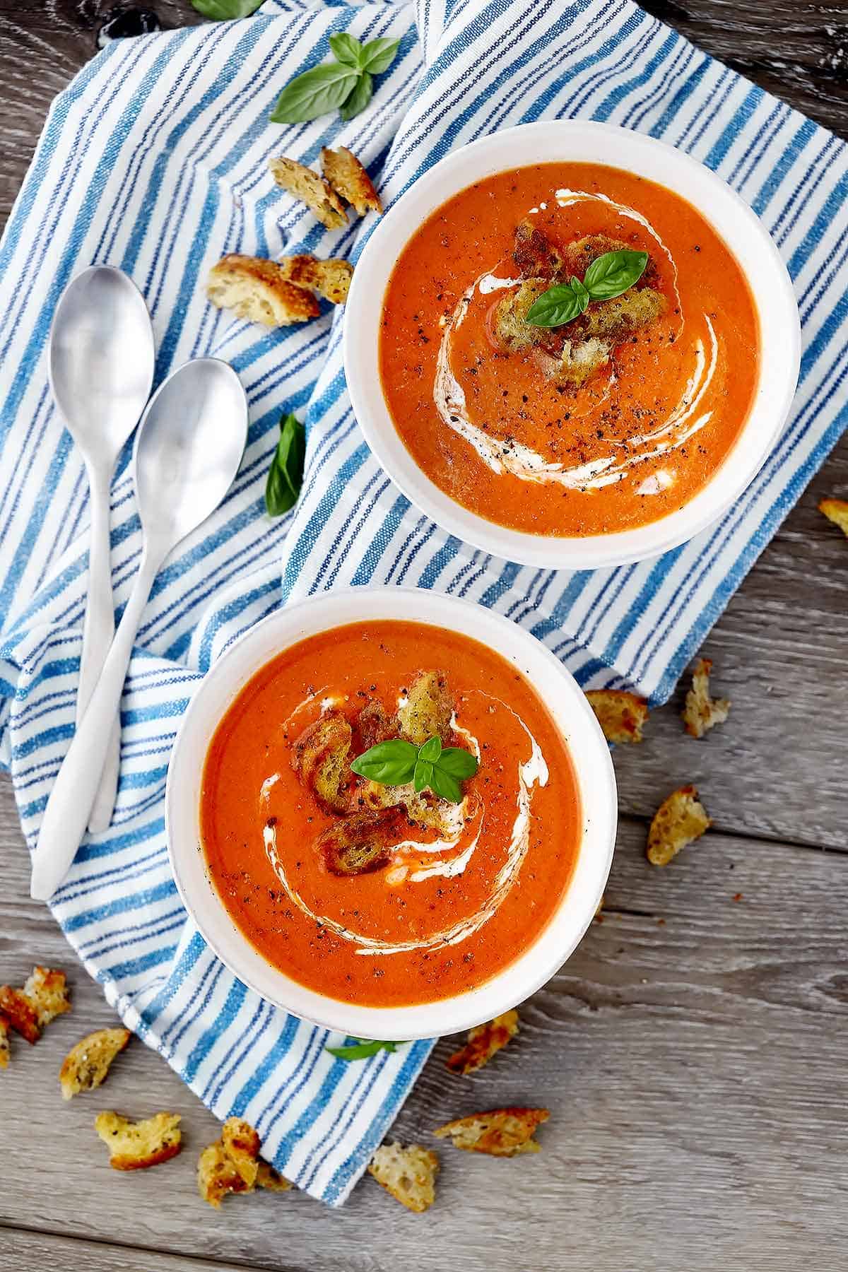 Overhead photo of two bowls of roasted red pepper and tomato soup on a towel with spoons