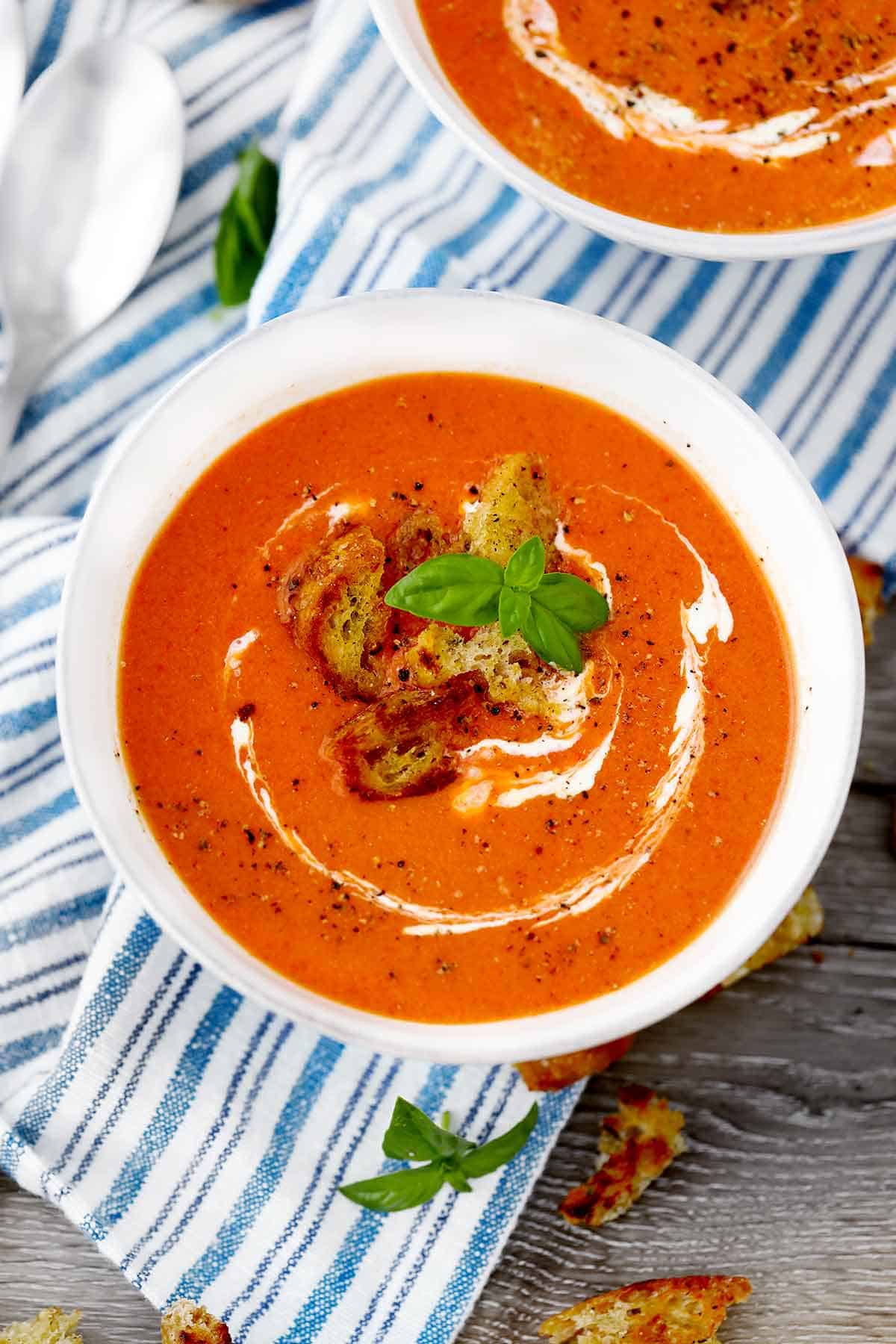 Close up photo of a bowl of roasted red pepper and tomato soup with basil leaves and croutons on top.