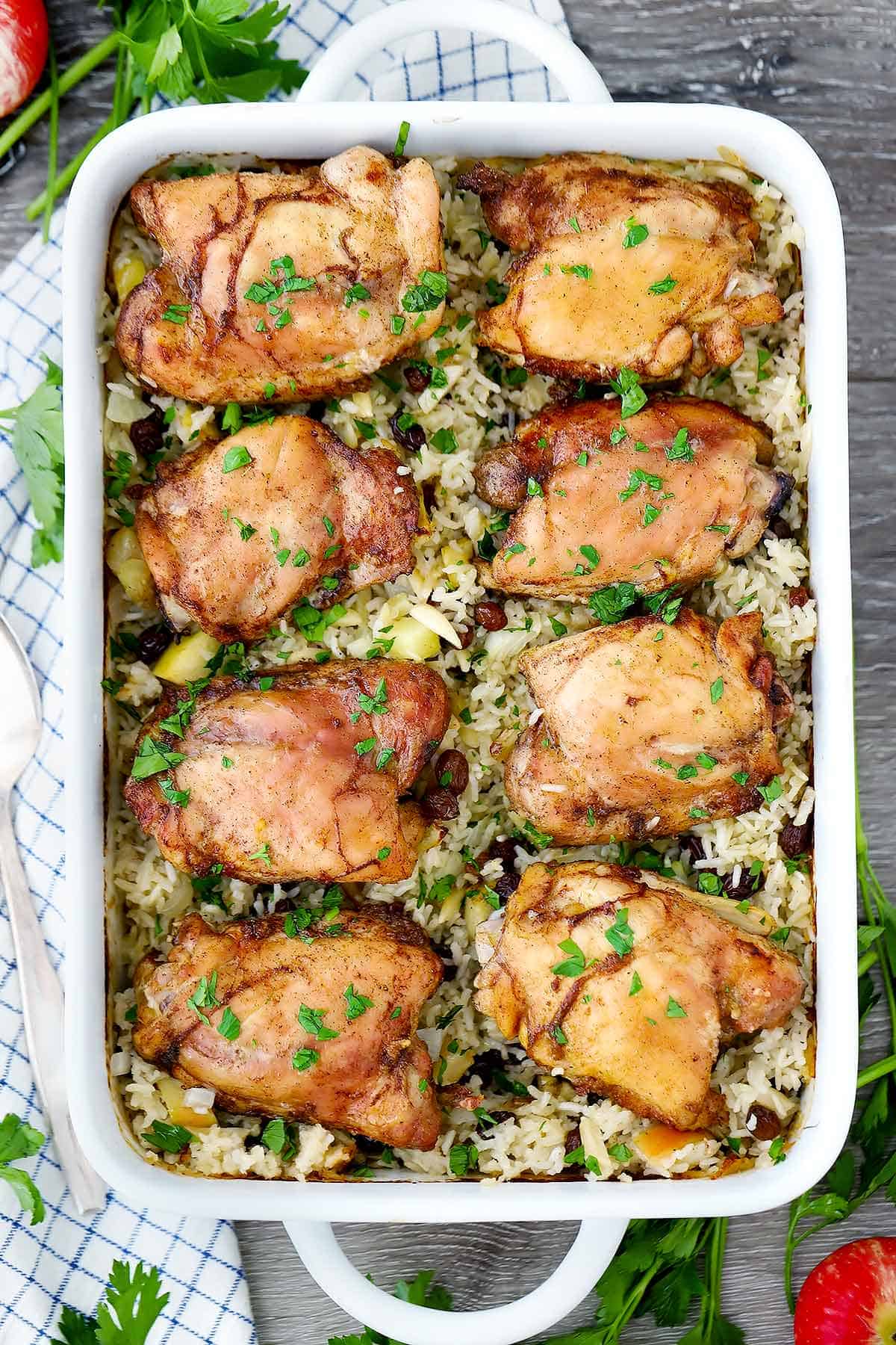 A casserole dish with chicken thighs baked on top of rice, apples, and raisins on a plaid towel.