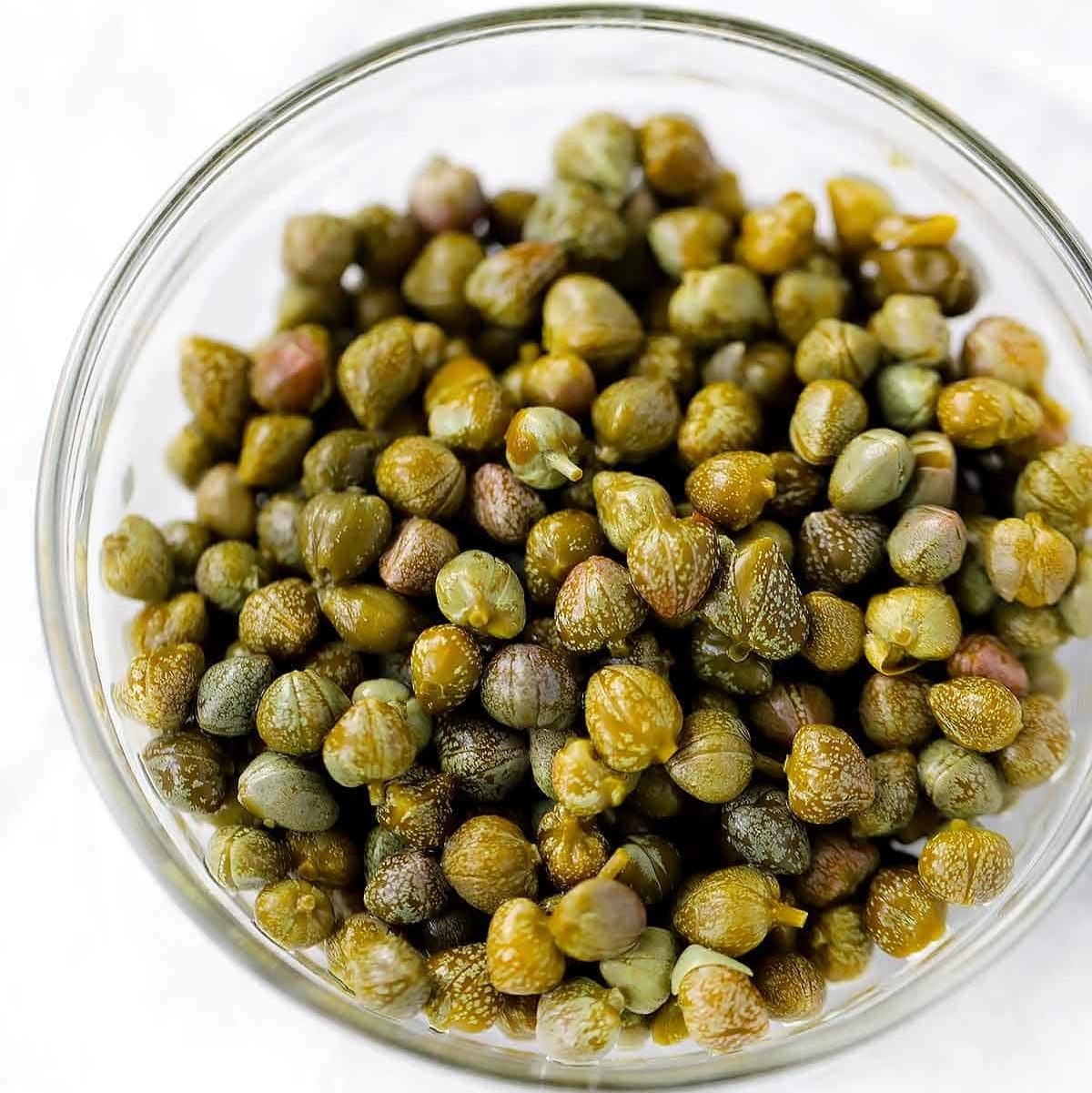 A close up photo of capers in a clear bowl with a white background.