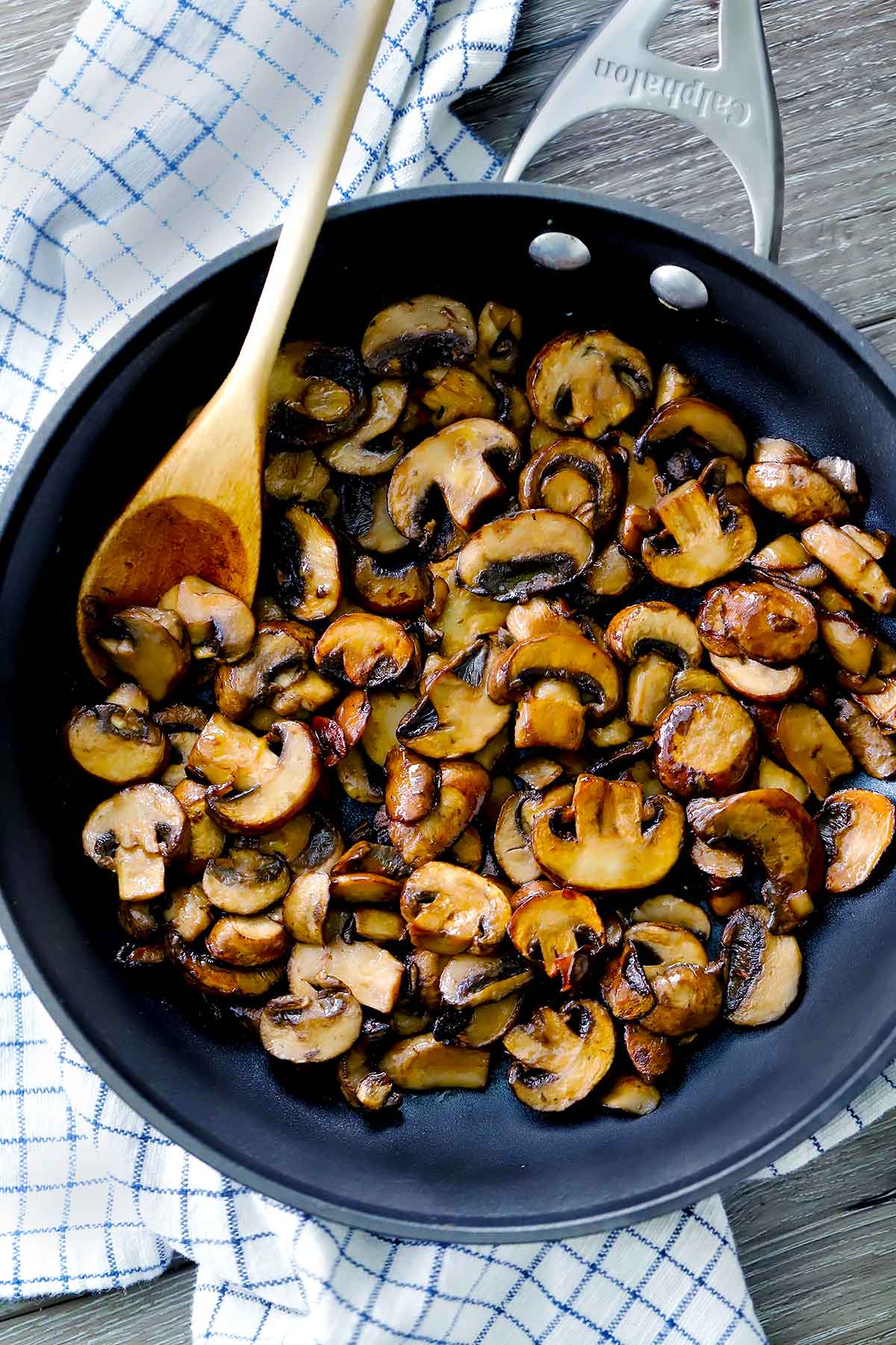 A skillet with sautéed mushrooms and a wooden spoon on a plaid towel.