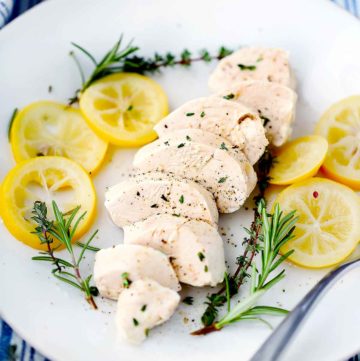 A sliced poached chicken breast with lemon, rosemary, and thyme around it on a white plate.