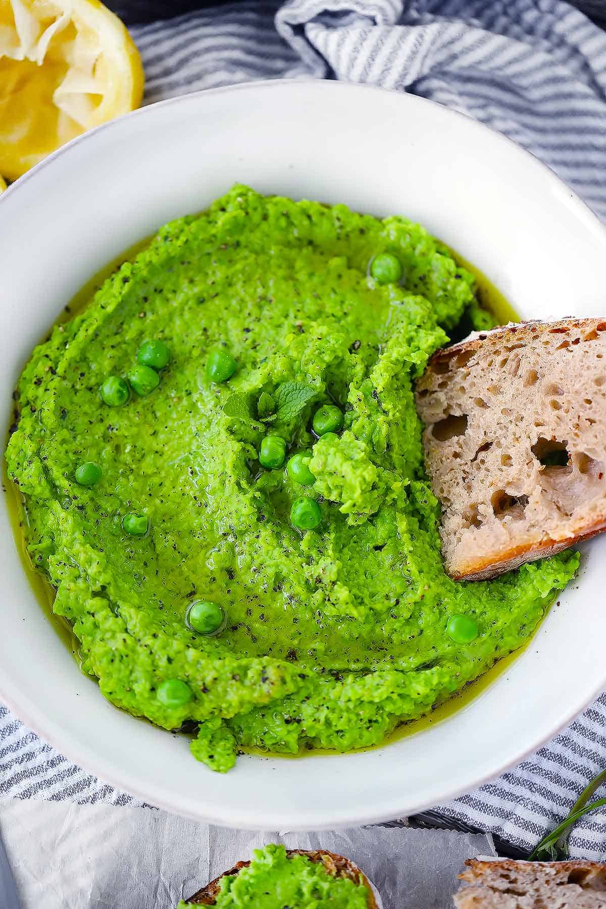 Overhead photo of pea puree in a white bowl with a piece of sourdough bread dipped into it.
