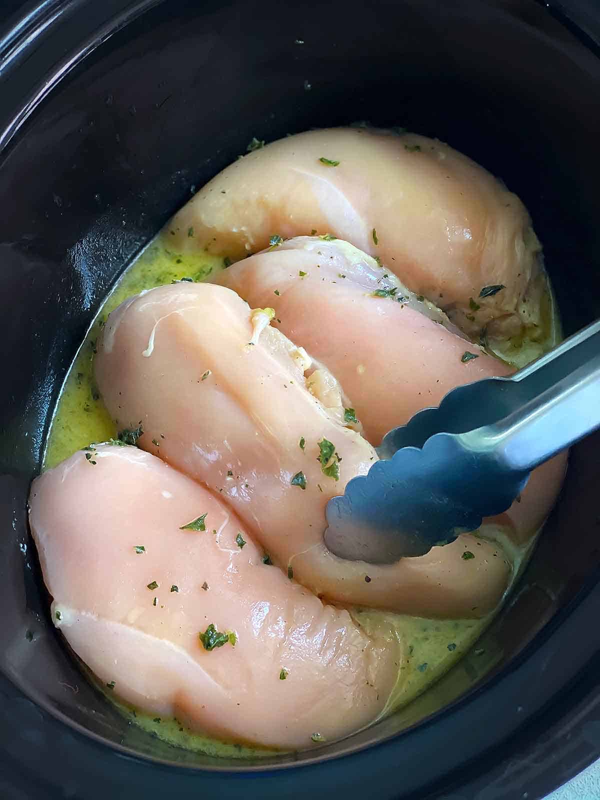 Adding raw chicken breasts to a slow cooker with tongs.