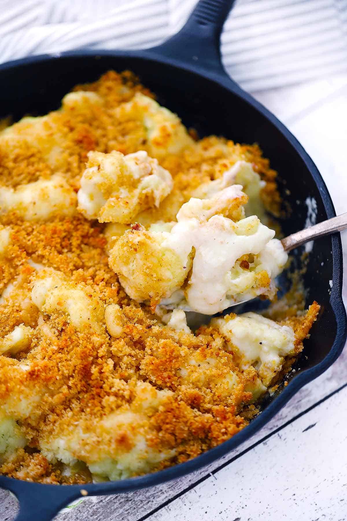 A serving spoon taking a scoop of cauliflower gratin out of a skillet.