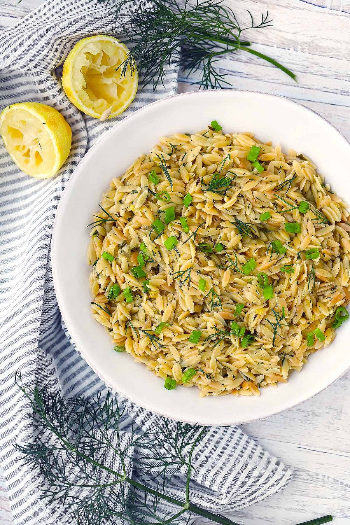 Overhead photo of orzo pilaf in a white bowl topped with green onions and dill with lemon on the side.