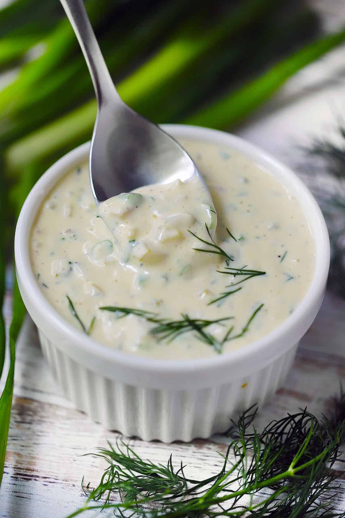 A white ramekin filled with homemade tartar sauce and a spoon scooping some out.