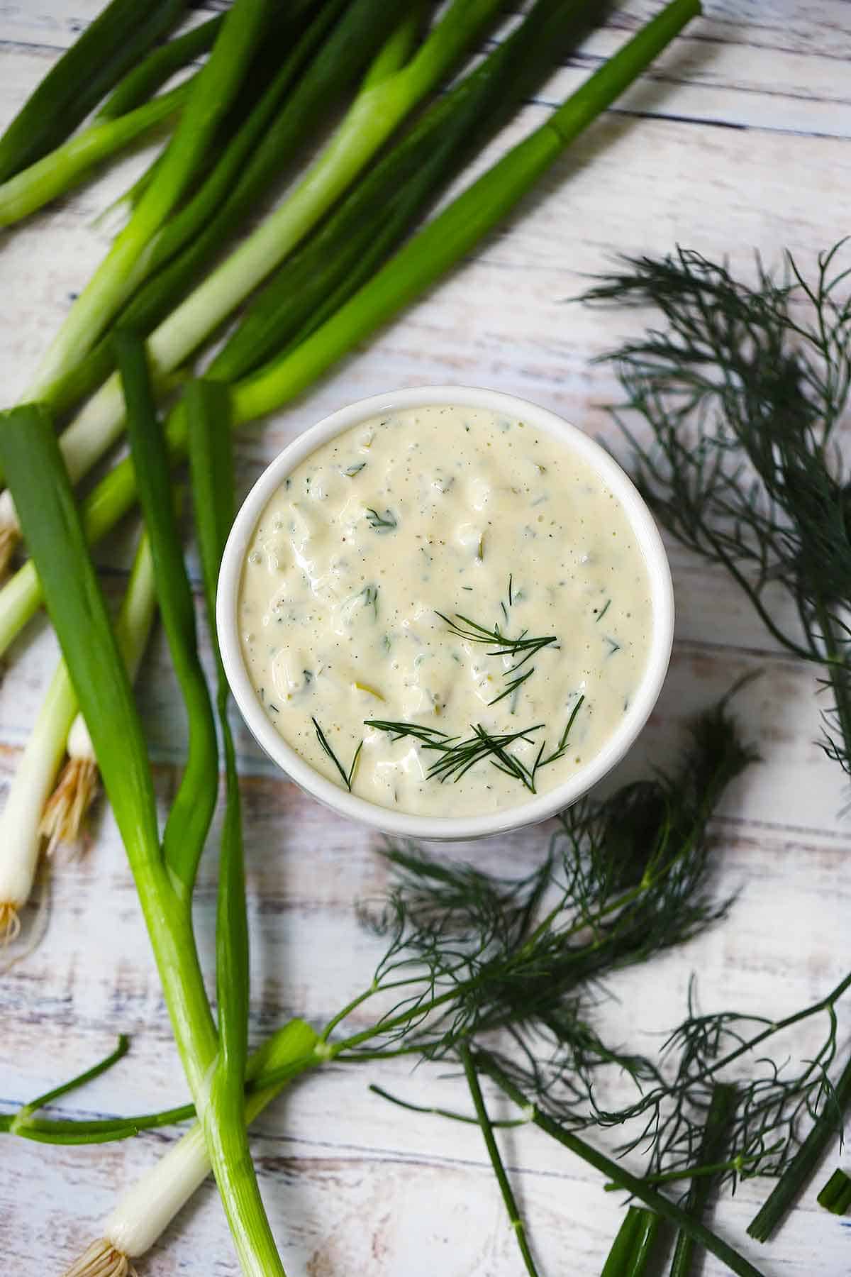 Overhead photo of a small bowl of tartar sauce surrounded by fresh dill and green onions.