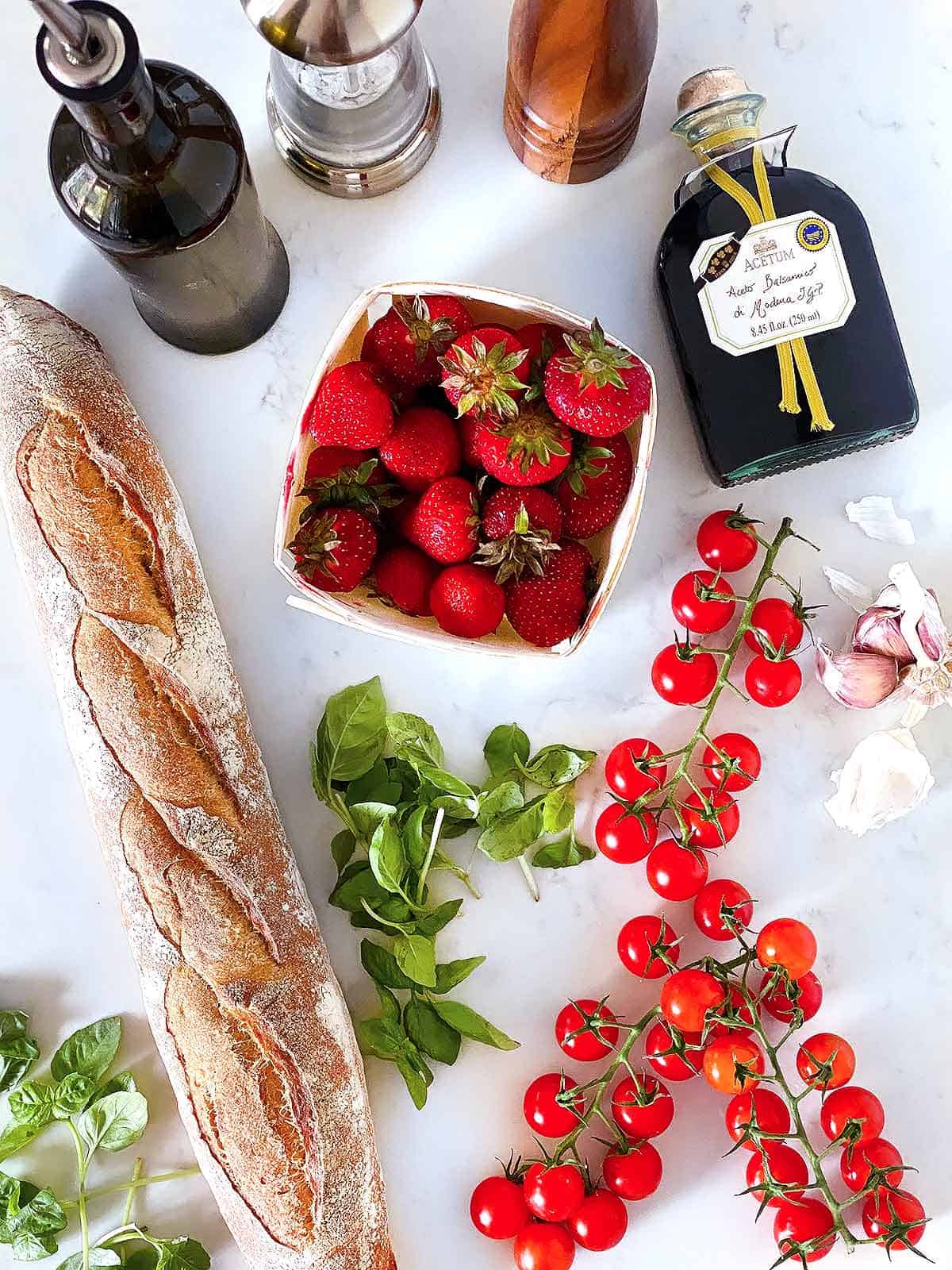 Overhead photo of a basket of strawberries, a baguette, fresh basil, cherry tomatoes, balsamic vinegar, oil, salt, and pepper.