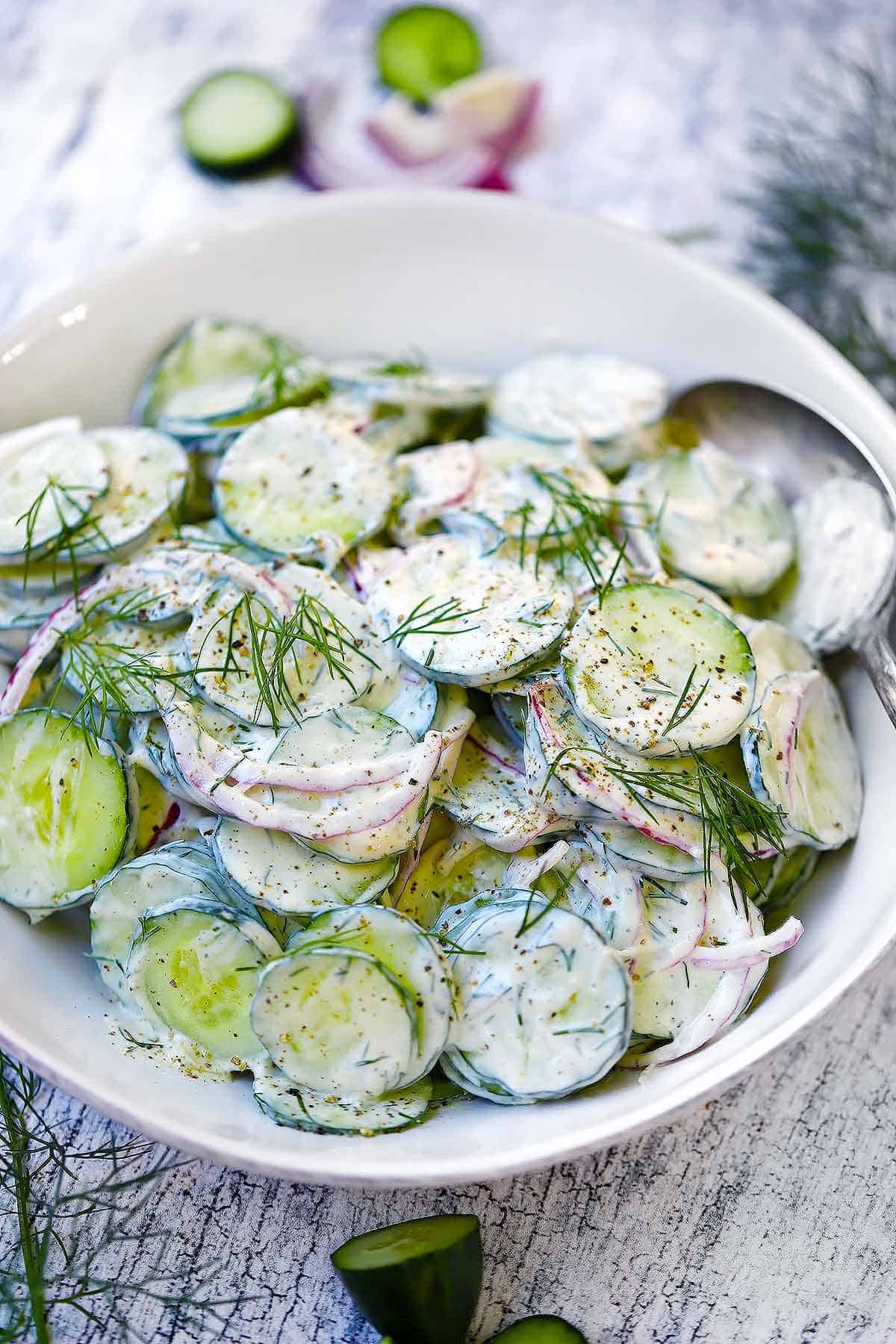 A white bowl with creamy cucumber salad in it and a spoon on the side.