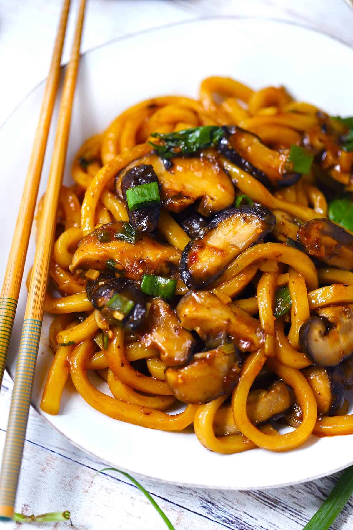 A white plate with udon noodles and shiitake mushrooms with green onions and chopsticks.