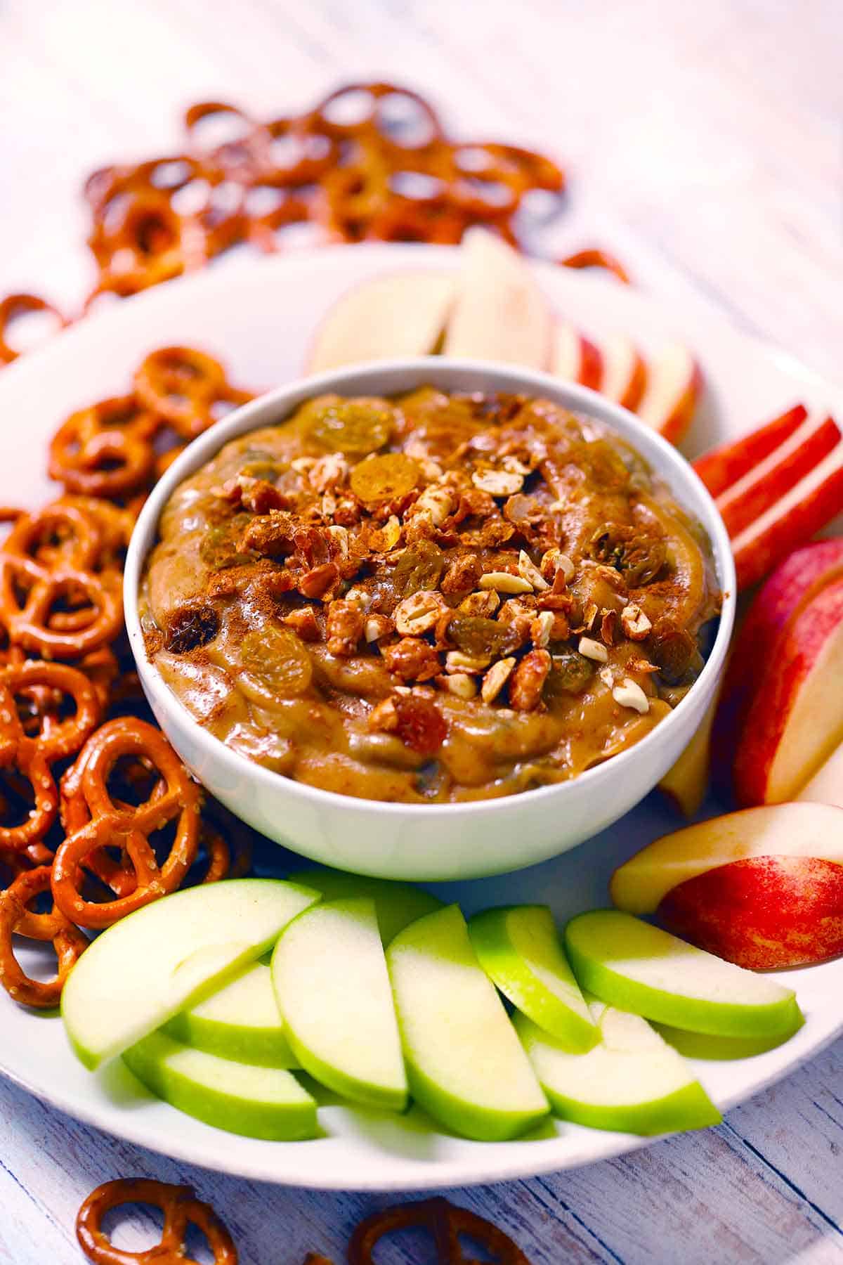 White bowl of peanut butter dip on a plate with apple slices and pretzels.